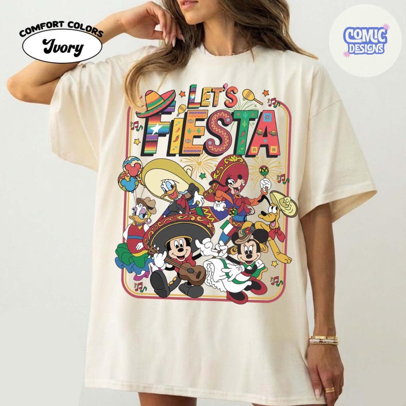 May include: Ivory-colored t-shirt with a colorful graphic featuring Disney characters in festive attire. The design includes Mickey Mouse playing a guitar, and the words "Let's Fiesta" in a vibrant, outlined font. The shirt has a relaxed fit.