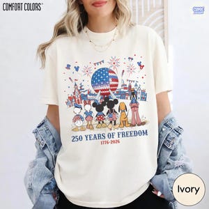May include: An ivory t-shirt featuring a graphic of cartoon characters standing before a large geodesic sphere with a flag pattern. Fireworks and balloons surround the scene. Text reads 250 YEARS OF FREEDOM and 1776-2026 in blue and red.
