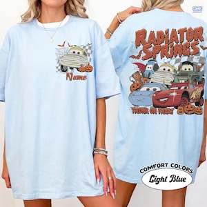 May include: A light blue t-shirt featuring a small graphic on the front chest of a mummy car with pumpkins and the word Name. The back displays a large graphic of multiple cartoon cars with Halloween themes, pumpkins, and the text Radiator Springs.