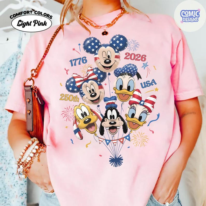 以下が含まれることがあります： ライトピンクのComfort Colors Tシャツ。ミッキー、ミニー、ドナルド、デイジー、グーフィー、プルートが愛国的なデザインの風船として描かれたグラフィック。Tシャツには「1776」、「2026」、「250th」、「USA」の文字が含まれています。