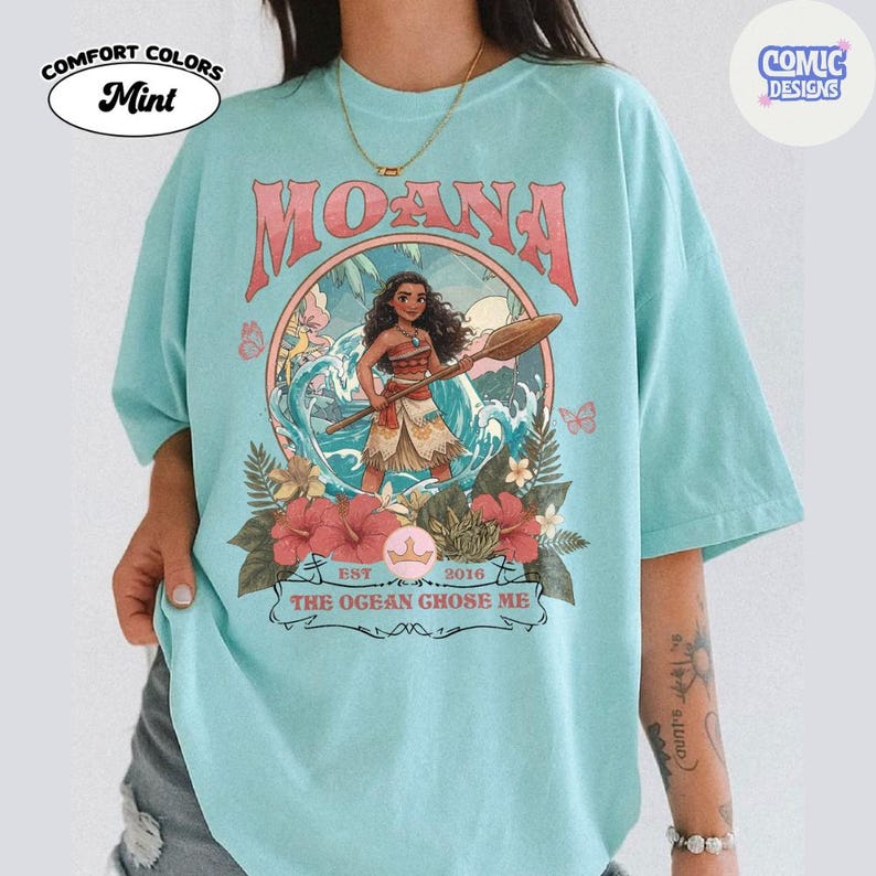 May include: A mint green t-shirt featuring a graphic print of the character Moana holding an oar, surrounded by ocean waves, tropical flowers, and foliage. The text reads MOANA and THE OGEAN GHOSE ME, with a small crown icon and EST 2016.