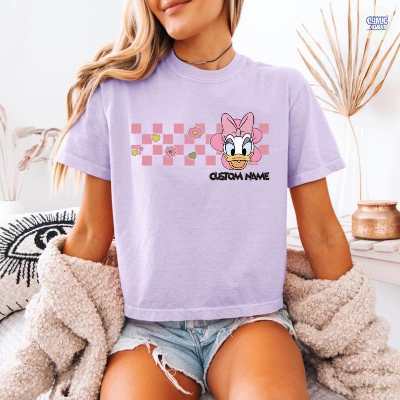 May include: A light purple t-shirt featuring a cartoon Daisy Duck design with a pink bow and a checkered pattern. The text "CUSTOM NAME" is printed below the character. The shirt is short-sleeved.