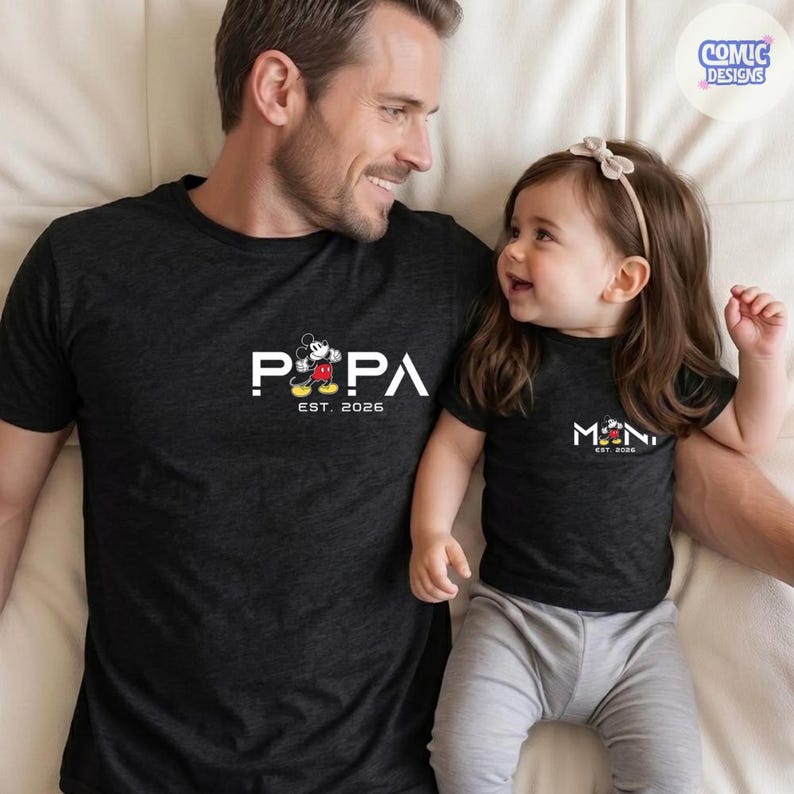 May include: Matching black t-shirts with white text and a Mickey Mouse graphic. One shirt says "PAPA EST. 2026" and the other says "MINI EST. 2026". The shirts are worn by a man and a child.