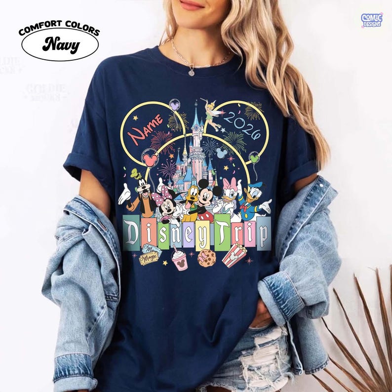 May include: Navy blue t-shirt with a colorful Disney Trip design. The design includes Disney characters, a castle, fireworks, and the year 2020. The shirt also features the word "Name" and the Comfort Colors logo.