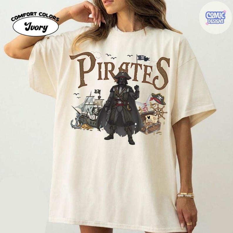 May include: Ivory-colored t-shirt with a "Pirates" graphic. The design features a pirate ship, treasure chest, and a character in a black outfit. The shirt has the words "Comfort Colors" and "Ivory" printed on it.