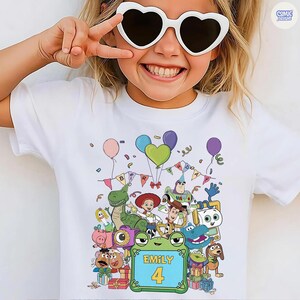 May include: A white short-sleeved t-shirt featuring a colorful graphic print of various animated toy characters, balloons, and birthday bunting. The center of the design includes a green frog-shaped frame with the text Emily and the number 4.