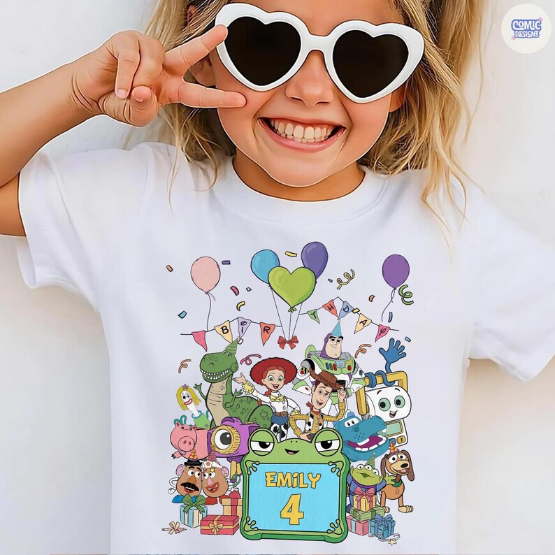 May include: A white short-sleeved t-shirt featuring a colorful graphic print of various animated toy characters, balloons, and birthday bunting. The center of the design includes a green frog-shaped frame with the text Emily and the number 4.