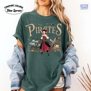 May include: A teal Comfort Colors t-shirt with the word "Pirates" in a vintage font. The shirt features a pirate-themed graphic with a woman in a red and black dress, a pirate ship, and a treasure chest. The text "Blue Spruce" is also visible.