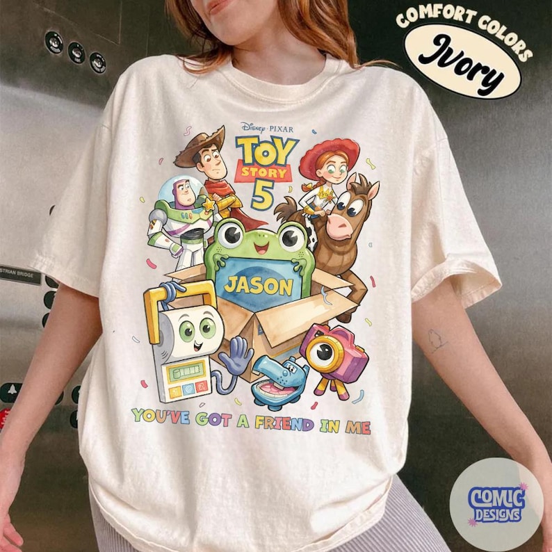 May include: An ivory t-shirt featuring a Toy Story 5 graphic with characters including Woody, Buzz Lightyear, Jessie, and Bullseye. A green frog sits in a cardboard box labeled Jason. The text reads Toy Story 5 and You've Got A Friend In Me.