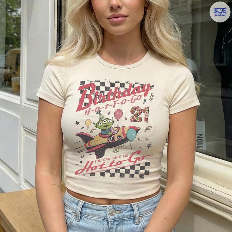 May include: Cream-colored, short-sleeved crop top with a graphic featuring a rocket ship and the text "Birthday H-O-T-T-O-GO 21". The design includes an alien character and balloons. The shirt is paired with blue jeans.