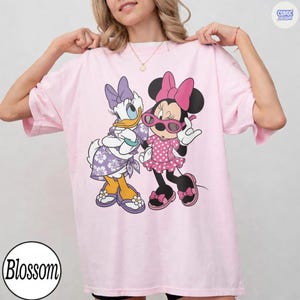 Puede incluir: Una camiseta rosa claro con un estampado gr&aacute;fico de Daisy Duck y Minnie Mouse. Daisy lleva un vestido floral morado y un lazo, mientras que Minnie lleva un vestido rosa de lunares, un lazo rosa y gafas de sol rosas. Cuello redondo.