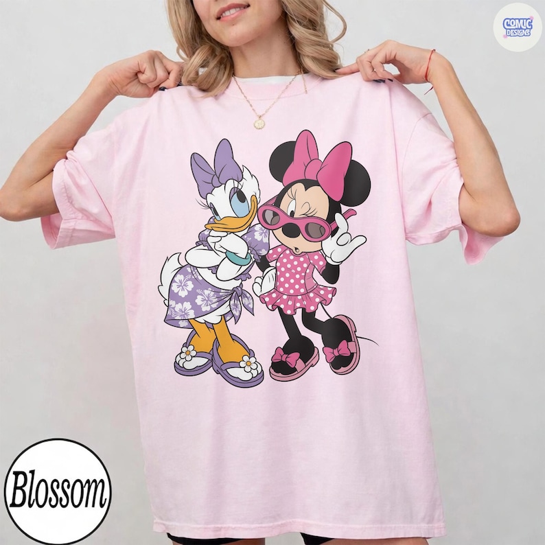 Puede incluir: Una camiseta rosa claro con un estampado gr&aacute;fico de Daisy Duck y Minnie Mouse. Daisy lleva un vestido floral morado y un lazo, mientras que Minnie lleva un vestido rosa de lunares, un lazo rosa y gafas de sol rosas. Cuello redondo.