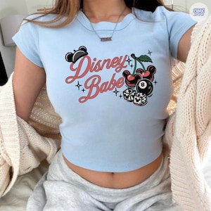 May include: Pale blue crop top featuring the text "Disney Babe" in red script, decorated with black Mickey Mouse ears, cherries, dice, and an 8-ball graphic. The top has short sleeves and a scoop neckline.