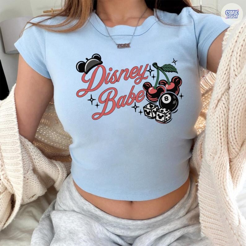 May include: Pale blue crop top featuring the text "Disney Babe" in red script, decorated with black Mickey Mouse ears, cherries, dice, and an 8-ball graphic. The top has short sleeves and a scoop neckline.