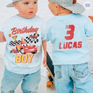 Puede incluir: Una camiseta azul claro con tem&aacute;tica de cumplea&ntilde;os. La parte delantera presenta un coche de carreras rojo, el texto "Birthday Boy" y otros gr&aacute;ficos. La parte trasera de la camiseta muestra el n&uacute;mero "3" y el nombre "LUCAS" en rojo.