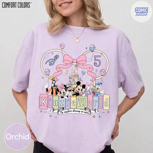 Op de afbeelding: Orchidee-kleurig T-shirt met een Disney-thema. Het ontwerp bevat Disney-personages, een kasteel, een grote roze strik en de tekst "Kinzie World". Het nummer "5" is ook aanwezig, samen met de zin "The happiest birthday on Earth".