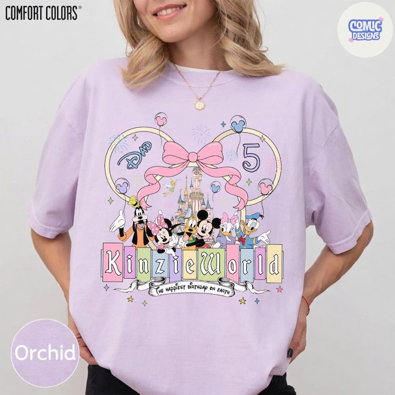 Op de afbeelding: Orchidee-kleurig T-shirt met een Disney-thema. Het ontwerp bevat Disney-personages, een kasteel, een grote roze strik en de tekst "Kinzie World". Het nummer "5" is ook aanwezig, samen met de zin "The happiest birthday on Earth".