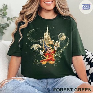 May include: Forest green t-shirt featuring a graphic of Mickey Mouse in a wizard costume with a castle and sparkling accents. The text "COMIC DESIGNS" is in a circle in the upper right corner. The words "FOREST GREEN" are at the bottom.