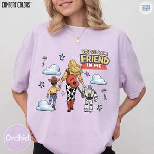 May include: A light purple t-shirt featuring a graphic of a person with long blonde hair holding hands with a cowboy figure and a space ranger figure. The design includes a guitar, clouds, stars, and the text "YOU'VE GOT A FRIEND IN ME".