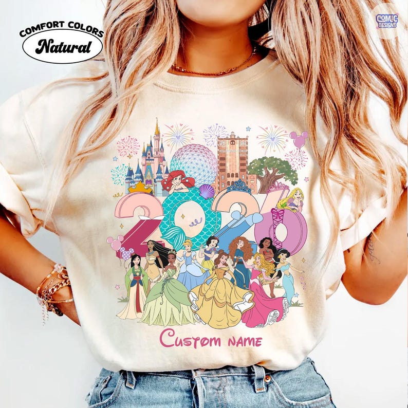 May include: A natural-colored t-shirt featuring a graphic print of the year 2024 with the numbers filled with patterns and surrounded by various animated princess characters, theme park landmarks, fireworks, and the text Custom Name at the bottom.