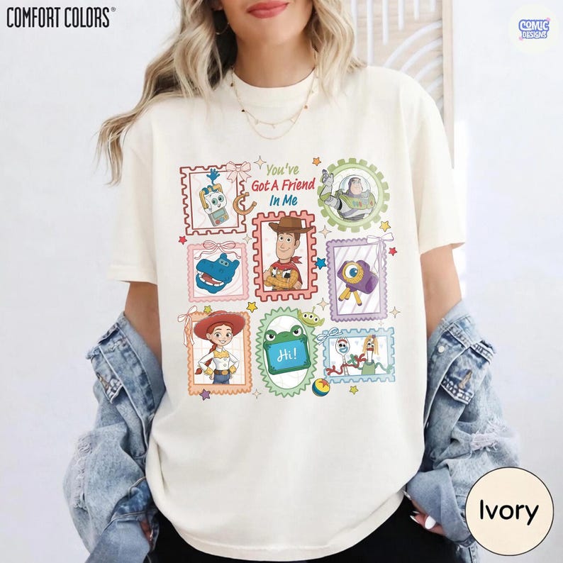 May include: An ivory t-shirt featuring a graphic print of various Toy Story characters arranged in colorful postage stamp-style frames. The text "You've Got A Friend In Me" is printed in the center, surrounded by illustrations of Woody, Buzz Lightyear, Jessie, and other characters.