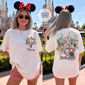 May include: An off-white t-shirt featuring a small graphic of Minnie Mouse and the word Mommy on the front left chest. The back displays a large, colorful illustration of Mickey, Minnie, Donald, and Pluto at Disneyland with the text Disneyland Est. 1955.