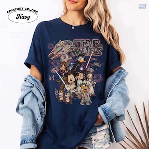 May include: Navy blue Comfort Colors t-shirt featuring a Star Wars graphic with various Disney characters, fireworks, and an American flag theme. The shirt has rolled-up sleeves and a crew neck.