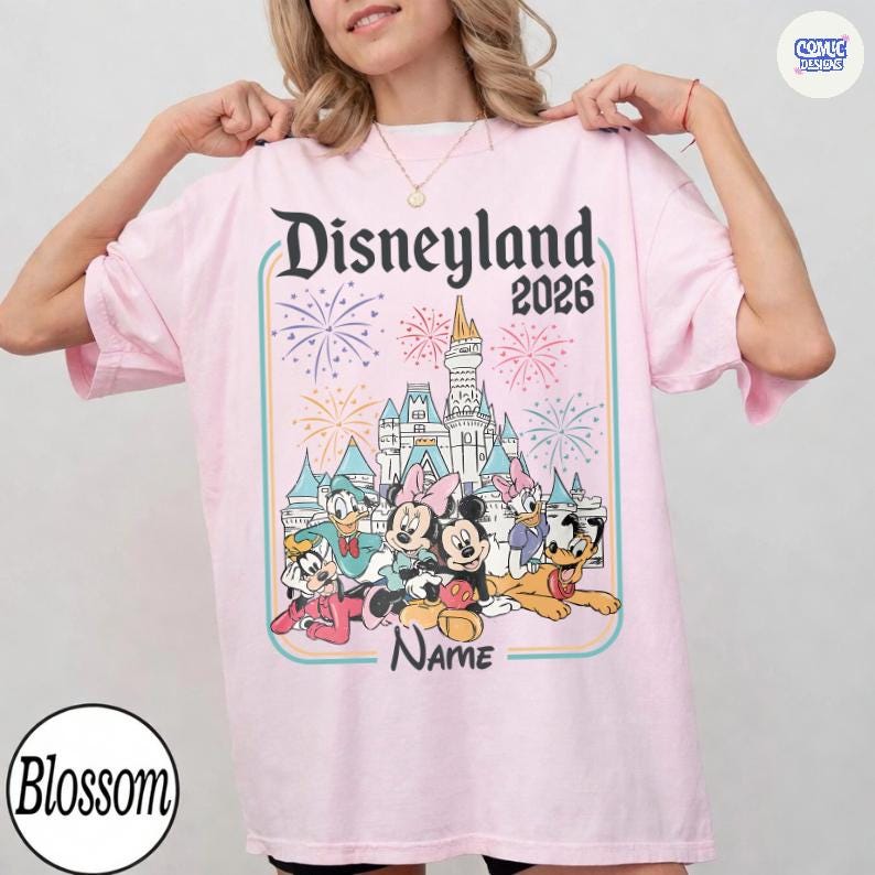 Op de afbeelding: Lichtroze t-shirt met het Disneyland-logo en het jaar 2026. Het ontwerp bevat een kasteel, vuurwerk en stripfiguren. Het woord "Name" is ook zichtbaar.
