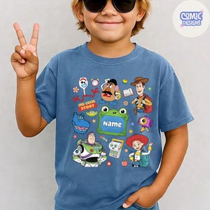 May include: A blue short-sleeved t-shirt featuring a colorful graphic print of various animated characters including a space ranger, a cowboy, a cowgirl, a potato figure, and a fork character. The design includes a green frame with the text Name.