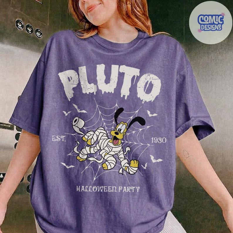 May include: A purple short-sleeve t-shirt featuring a graphic of Pluto wrapped in toilet paper like a mummy in front of a spider web. The text reads PLUTO, EST. 1930, and HALLOWEEN PARTY, accompanied by small bat illustrations.