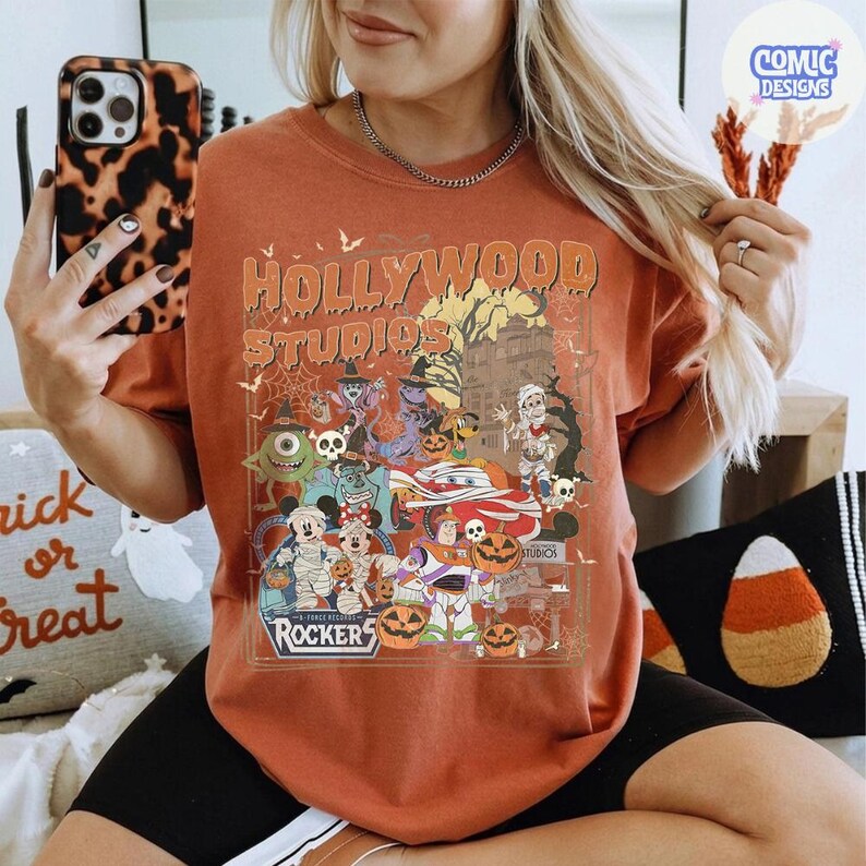 K&ouml;nnte beinhalten: Ein orangefarbenes T-Shirt mit einem Grafikdruck mit dem Text Hollywood Studios und Rockers. Das Design zeigt verschiedene Zeichentrickfiguren in Halloween-Kost&uuml;men, K&uuml;rbisse, Spinnennetze und die Silhouette eines Spukhauses.