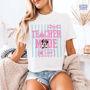 May include: White t-shirt with the text "TEACHER MODE OFF" in pink, with a Minnie Mouse graphic. The design includes a pink bow and light blue vertical stripes. The shirt is short-sleeved.