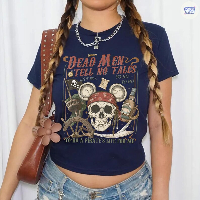以下が含まれることがあります： ネイビーブルーのTシャツで、ネズミの耳が付いた赤いバンダナを巻いたドクロのグラフィックが描かれています。テキストには「Dead Men Tell No Tales」「Est 1967」「Yo Ho Yo Ho」「Yo Ho A Pirate's Life For Me」と書かれています。デザインには海賊旗、ボトル、舵輪が含まれています。