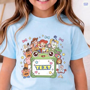 May include: A light blue t-shirt featuring a colorful graphic print of various animated characters surrounding a central green frog-shaped frame with the word TEXT printed in the center. The design includes stars, bows, and hearts.