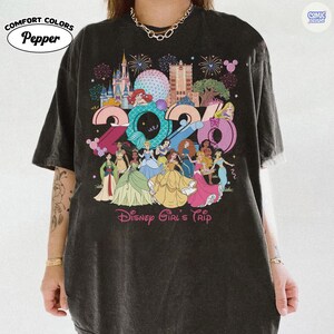 May include: A charcoal gray short-sleeve t-shirt featuring a large graphic print of the year 2024 with various animated princesses, castle silhouettes, and fireworks. The text Disney Girl's Trip is printed in pink at the bottom of the graphic design.