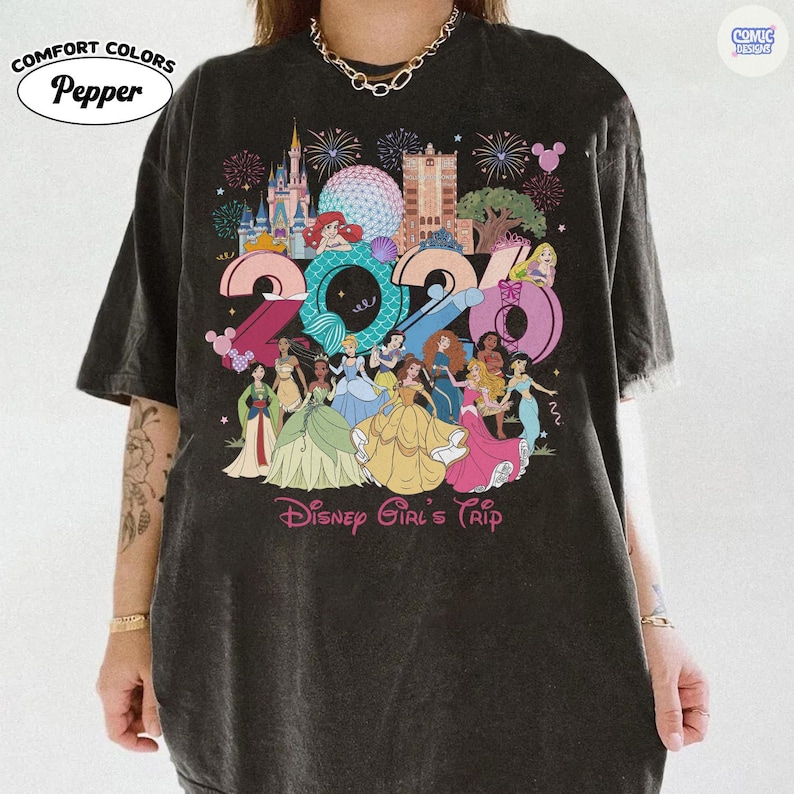 May include: A charcoal gray short-sleeve t-shirt featuring a large graphic print of the year 2024 with various animated princesses, castle silhouettes, and fireworks. The text Disney Girl's Trip is printed in pink at the bottom of the graphic design.