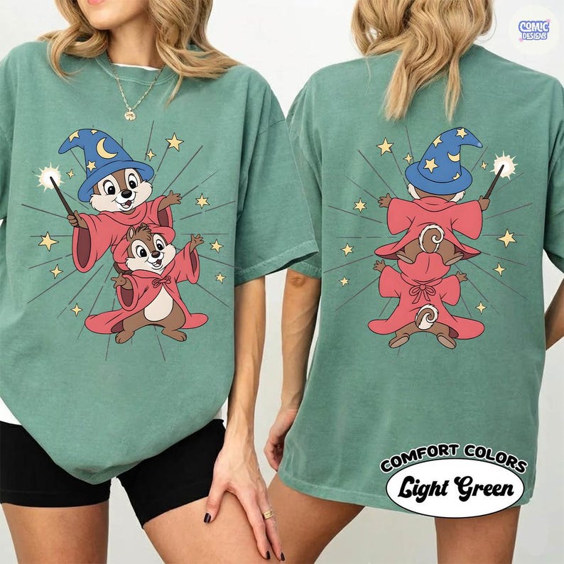 Puede incluir: Camiseta verde claro con una ilustraci&oacute;n de dibujos animados de dos ardillas vestidas de magos, una encima de la otra, con una varita m&aacute;gica y estrellas. La parte trasera de la camiseta tambi&eacute;n tiene el mismo dise&ntilde;o. El texto "COMFORT COLORS Light Green" est&aacute; impreso en la parte inferior derecha.