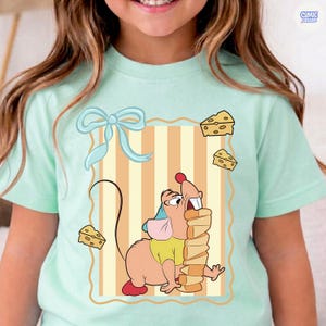 May include: A light green t-shirt featuring a cartoon illustration of a mouse with a stack of cheese. The mouse is wearing a yellow shirt and a blue hat. The design includes a light blue bow and cheese slices.