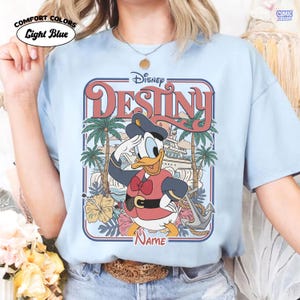 Op de afbeelding: Lichtblauw Comfort Colors T-shirt met een Disney-afbeelding. De afbeelding toont Donald Duck in een matrozenoutfit, het woord "DESTINY", palmbomen, een schip en het woord "NAME". Het shirt heeft de woorden "COMIC DESIGNS" en "Light Blue" erop gedrukt.