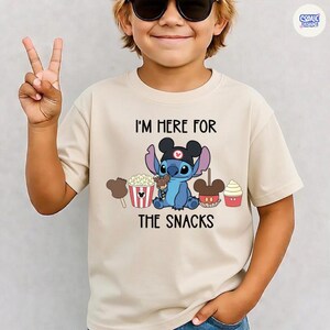 May include: Beige t-shirt featuring the character Stitch from Lilo & Stitch, wearing a black hat and surrounded by snacks. The text "I'M HERE FOR THE SNACKS" is printed above and below the character. The shirt is worn by a child.