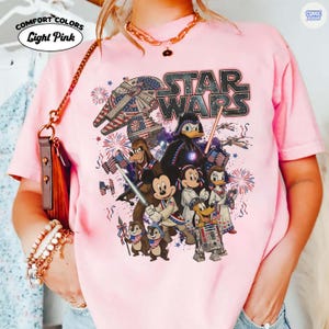 May include: Light pink Comfort Colors t-shirt featuring a Star Wars graphic with Disney characters. The design includes the Star Wars logo, the Millennium Falcon, Darth Vader, Mickey Mouse, Donald Duck, and fireworks. The shirt has the words "Light Pink" on it.
