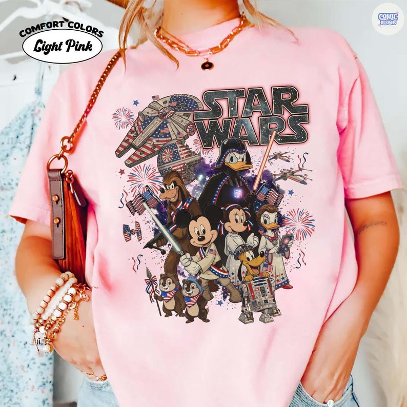 May include: Light pink Comfort Colors t-shirt featuring a Star Wars graphic with Disney characters. The design includes the Star Wars logo, the Millennium Falcon, Darth Vader, Mickey Mouse, Donald Duck, and fireworks. The shirt has the words "Light Pink" on it.