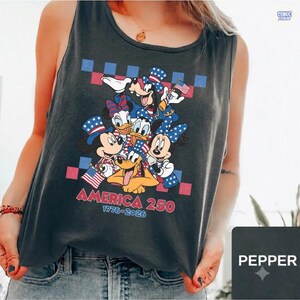 May include: A charcoal gray sleeveless tank top featuring a graphic print of cartoon characters wearing patriotic hats. The design includes the text AMERICA 250 and 1776-2026, set against a red and blue checkered background with American flag motifs.