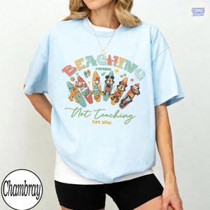 May include: Light blue t-shirt with a colorful graphic featuring cartoon characters on surfboards. The text reads "BEACHING #RETIRED Not Teaching EST. 2026". The shirt has a relaxed fit and short sleeves.