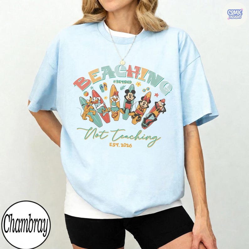 May include: Light blue t-shirt with a colorful graphic featuring cartoon characters on surfboards. The text reads "BEACHING #RETIRED Not Teaching EST. 2026". The shirt has a relaxed fit and short sleeves.