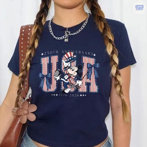 Op de afbeelding: Een marineblauw t-shirt met een afbeelding van Mickey Mouse in een patriottische hoed en outfit. De tekst luidt 250TH ANNIVERSARY USA 1776-2026, met rood-wit gestreepte letters en blauwe stippen-strikken aan de zijkanten van het grafische ontwerp.