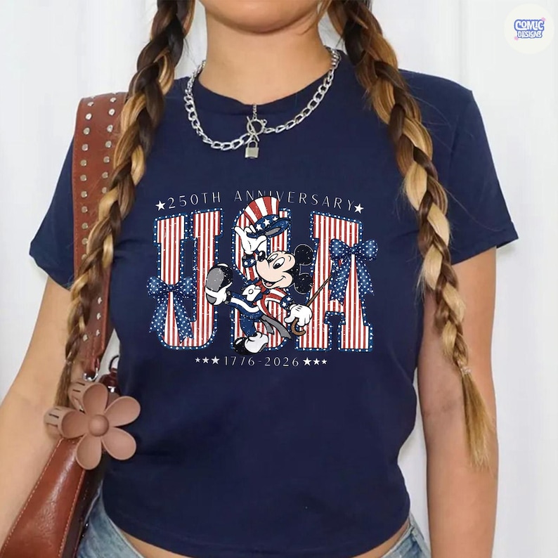 Op de afbeelding: Een marineblauw t-shirt met een afbeelding van Mickey Mouse in een patriottische hoed en outfit. De tekst luidt 250TH ANNIVERSARY USA 1776-2026, met rood-wit gestreepte letters en blauwe stippen-strikken aan de zijkanten van het grafische ontwerp.