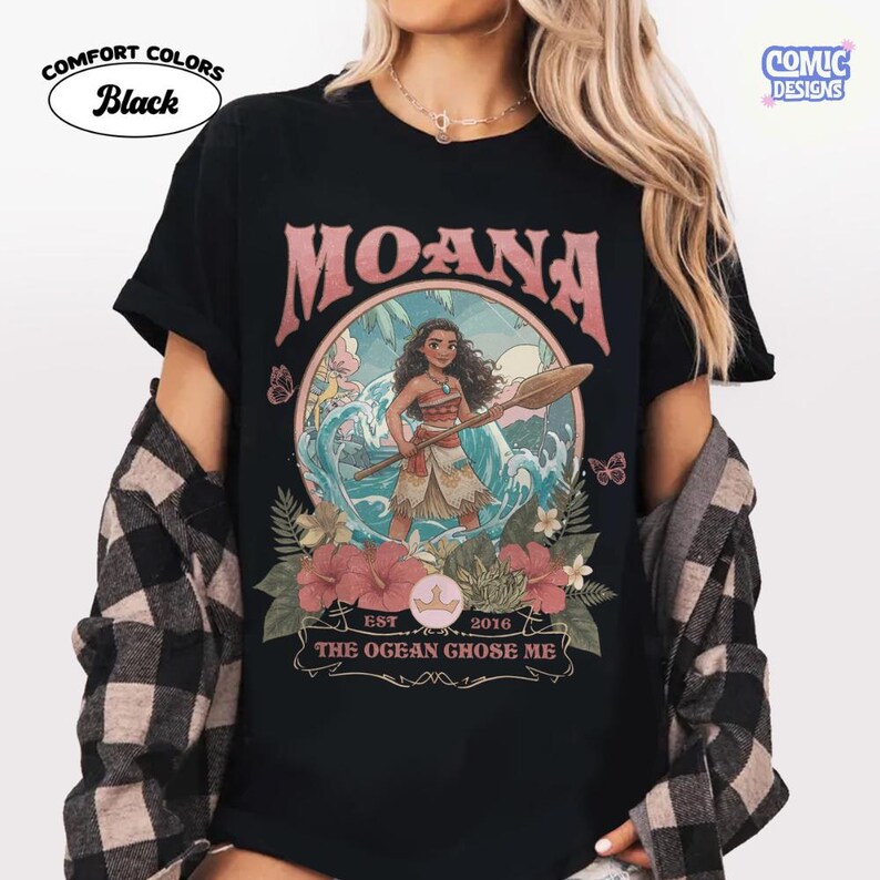 May include: A black short-sleeve t-shirt featuring a graphic of Moana holding an oar within a circular frame. The design includes tropical flowers, waves, and the text "MOANA" at the top and "EST 2016 THE OGEAN GHOSE ME" at the bottom in a vintage font.