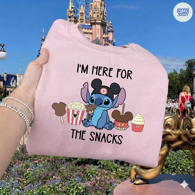 May include: A light pink sweatshirt featuring a cartoon Stitch character wearing a Mickey Mouse hat and surrounded by snack-themed graphics. The text "I'M HERE FOR THE SNACKS" is printed above and below the character.