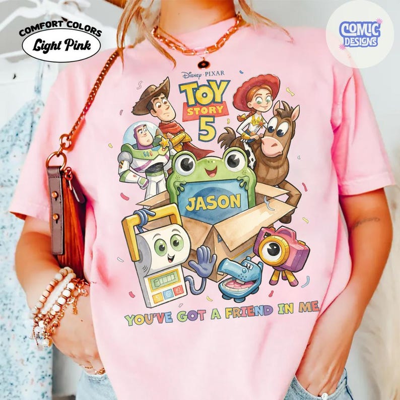 May include: A light pink t-shirt featuring a Toy Story 5 graphic with characters including Woody, Buzz Lightyear, Jessie, Bullseye, and a green frog in a box labeled Jason. The text reads Toy Story 5 and You've Got A Friend In Me.
